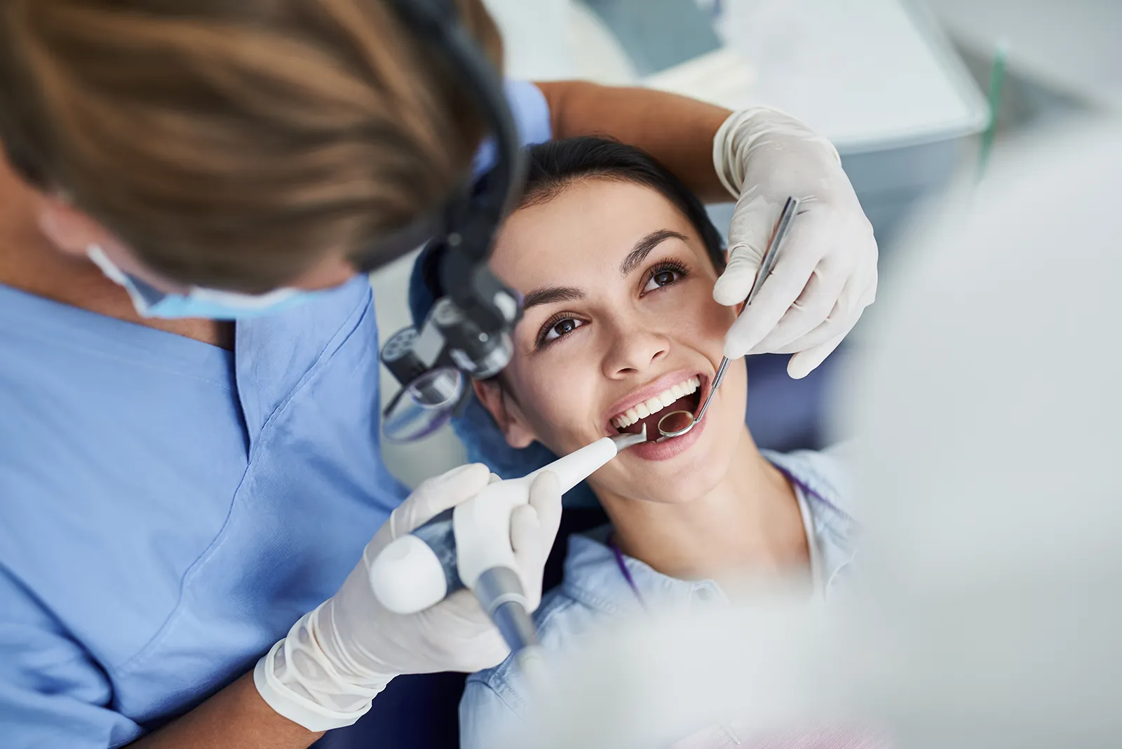 A dental hygienist in blue scrubs and magnifying loupes cleaning the teeth of a smiling patient in a bright treatment room.